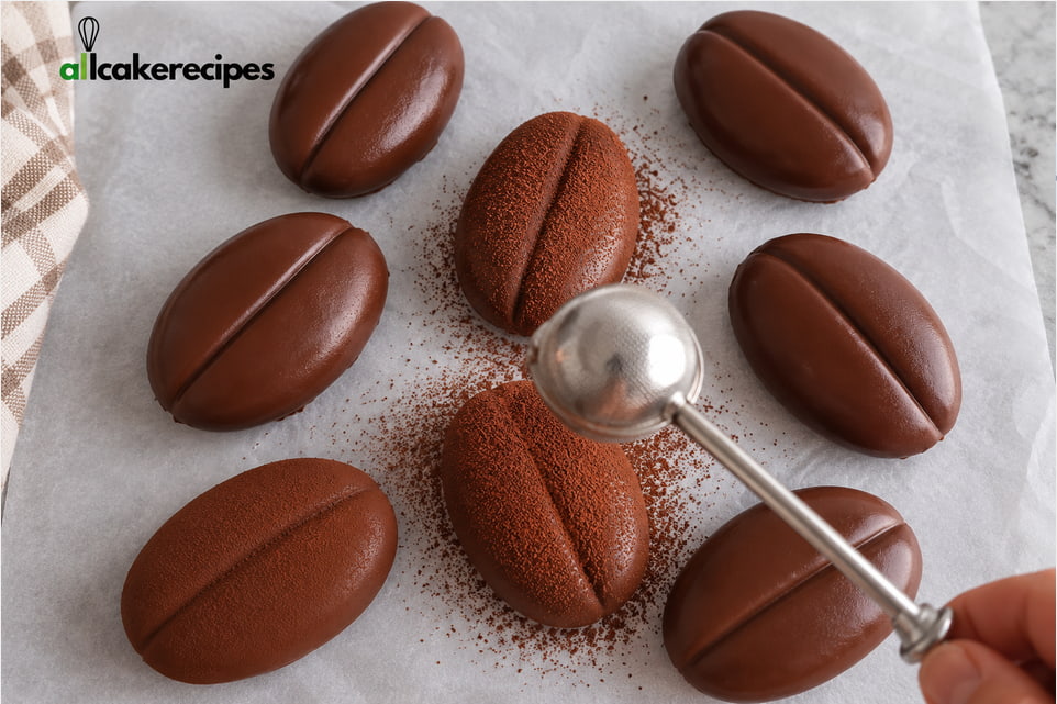 Dusting chocolate-coated coffee bean tiramisu desserts with unsweetened cocoa powder using a small sieve.