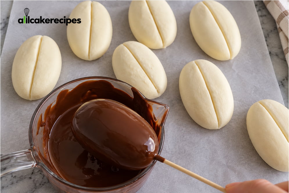 Dipping a coffee bean-shaped tiramisu dessert into melted dark chocolate for a smooth chocolate coating.